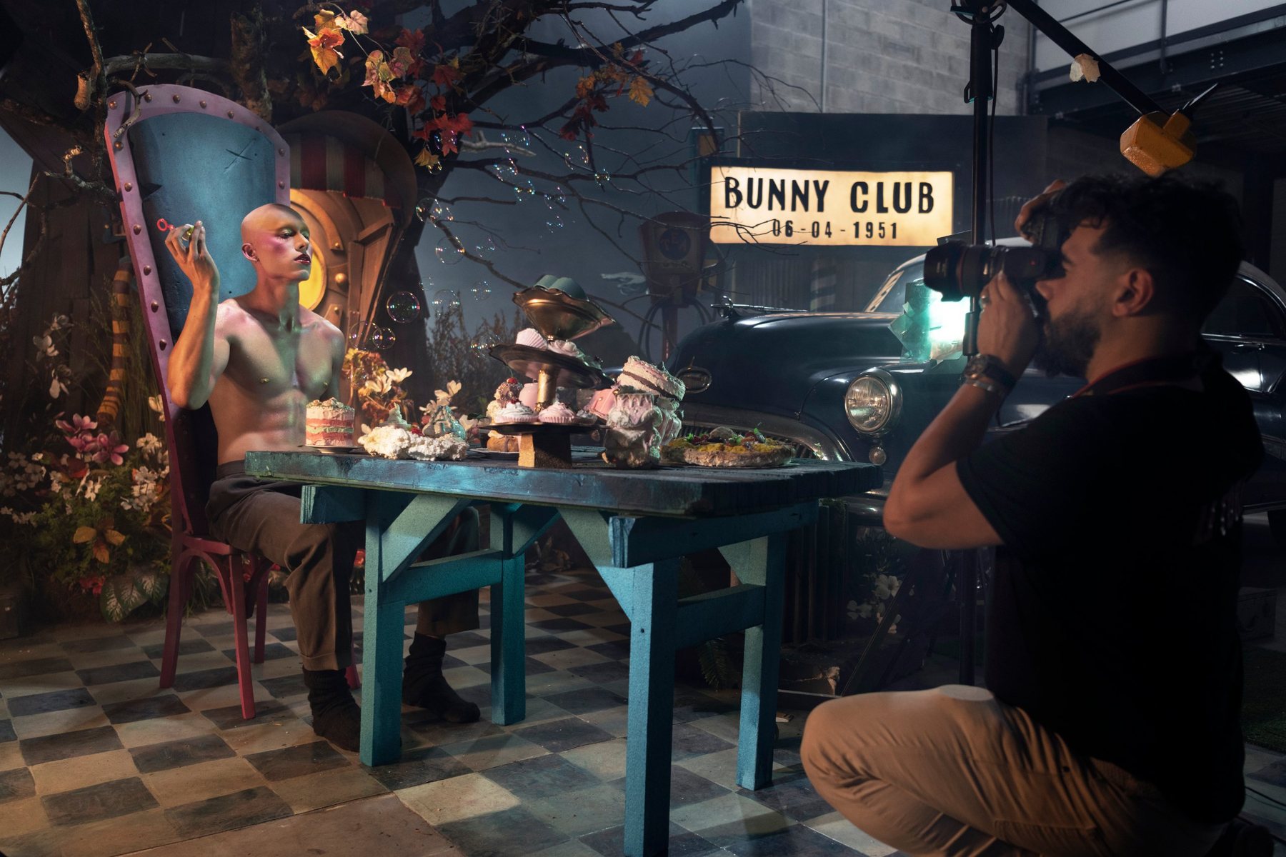 Un photographe à genoux cadre un modèle torse nu attablé devant des gâteaux, sous l’enseigne lumineuse « Bunny Club ».