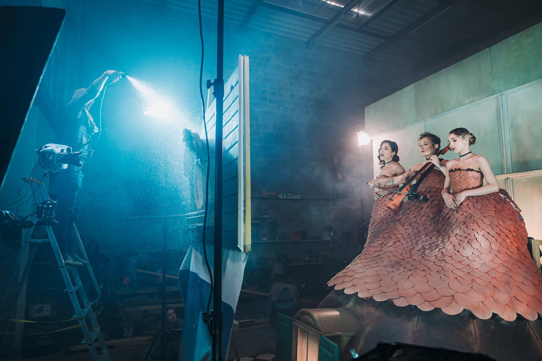 Vue large d’un tournage sous la pluie : un technicien arrose le décor depuis un escabeau, pendant que trois musiciennes en robes à écailles posent sur un toit construit dans l’atelier.