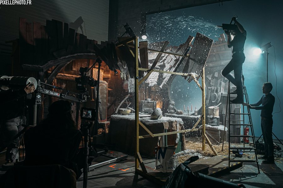 Tournage dans une cabane en bois sous une neige artificielle : un technicien perché sur un escabeau souffle la neige, une caméra sur grue filme la scène.