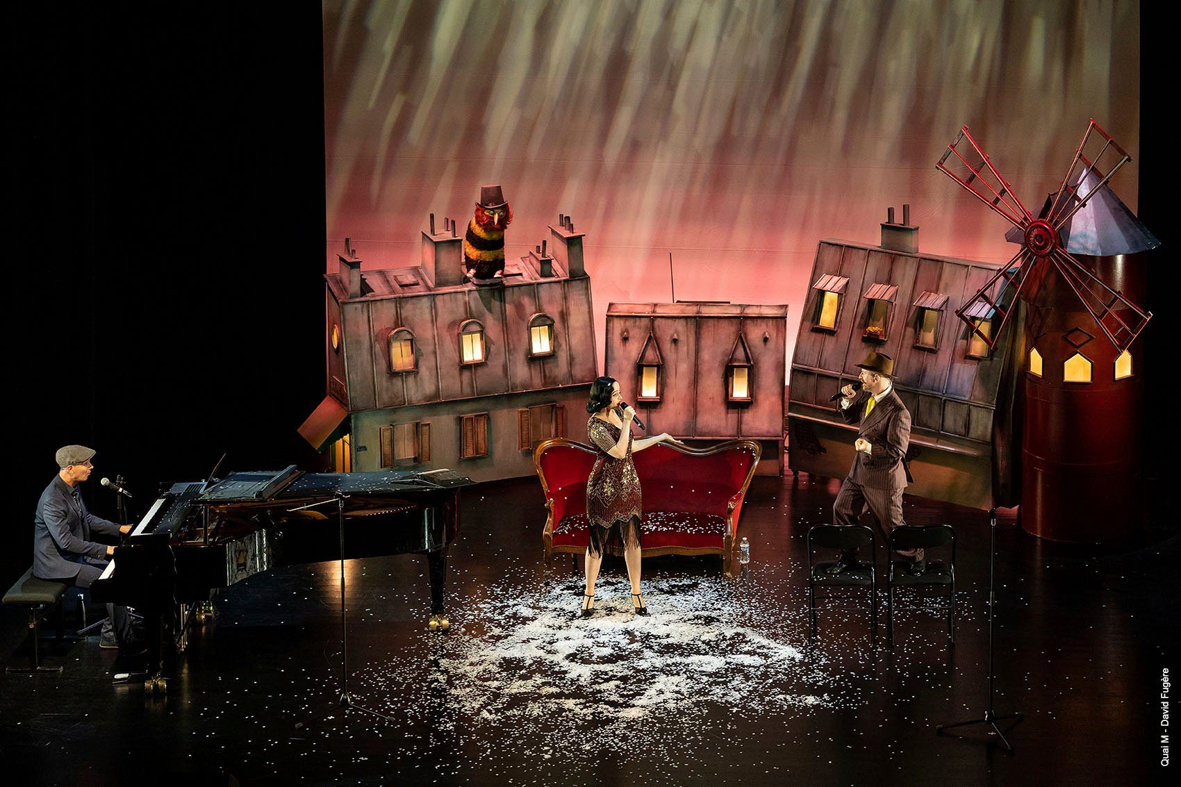 Spectacle dans un décor de toits de Paris penchés et de moulin rouge : une chanteuse près d’un canapé de velours, un chanteur et un pianiste, sous une neige de confettis.