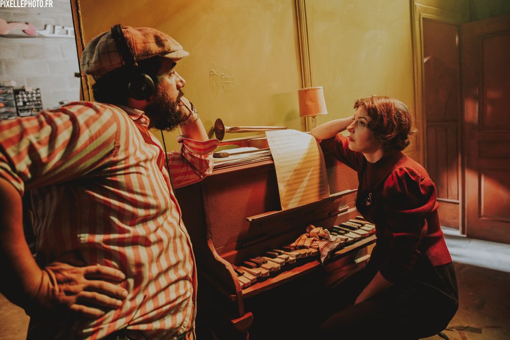 Le Turk, casque sur les oreilles, et Laura, accoudée à un vieux piano, sur un plateau éclairé.