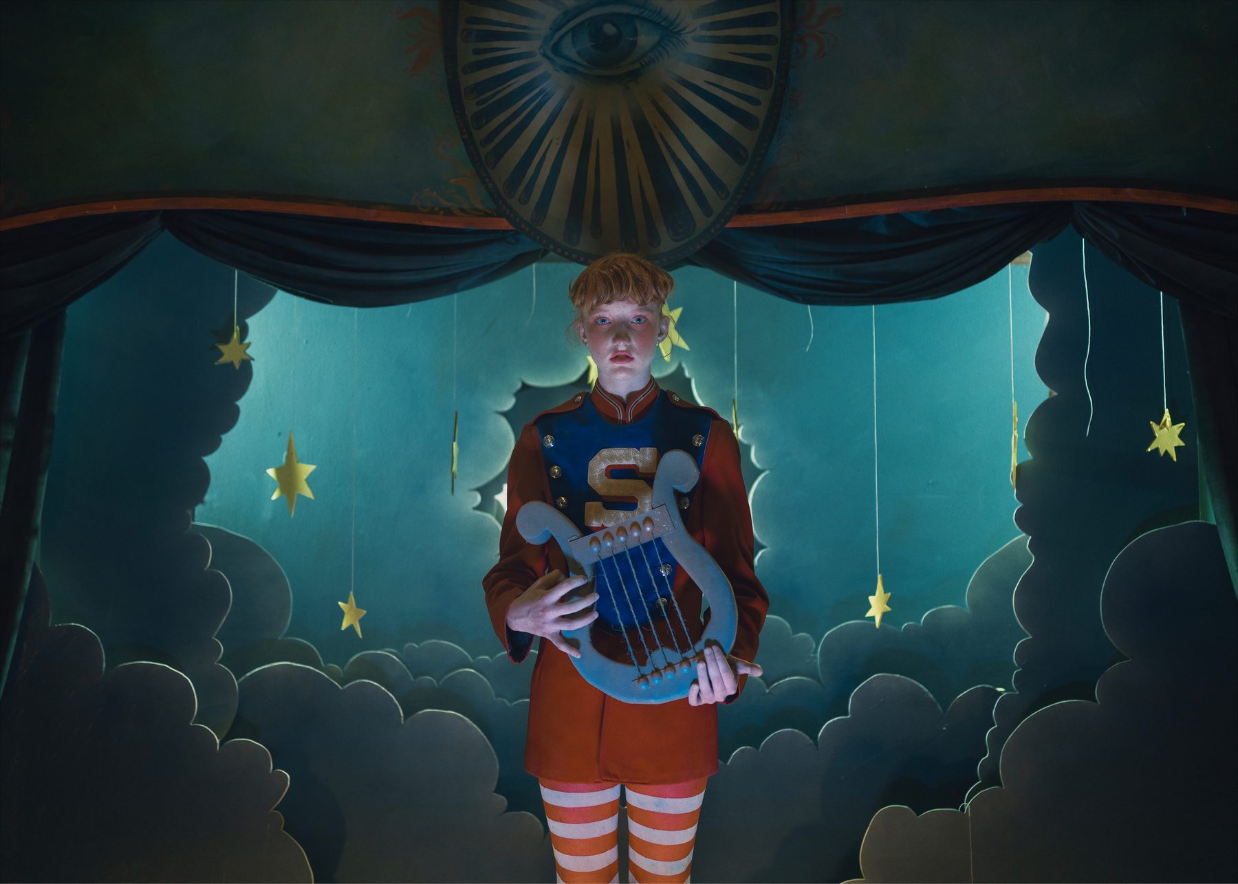 Un musicien en uniforme joue de la lyre sur une petite scène de nuages en carton et d’étoiles suspendues.