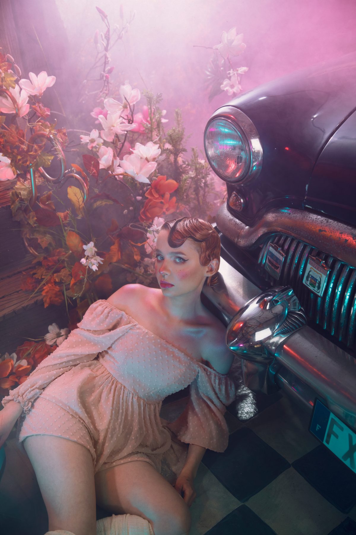 Une femme en robe pâle allongée contre une voiture ancienne envahie de fleurs roses.